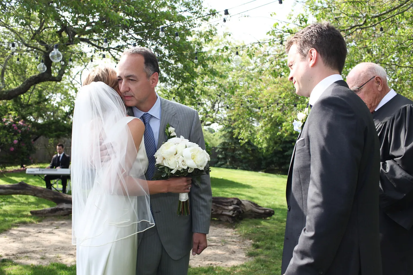 An older man in grey hugs the bride on the lawn as the groom and officiant wait