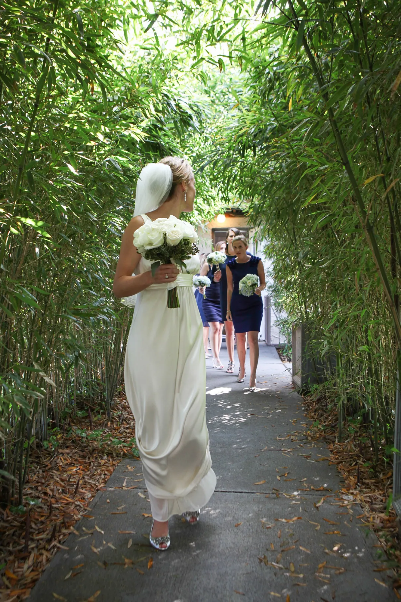 The bride walks the bamboo-lined aisle holding a white bouquet