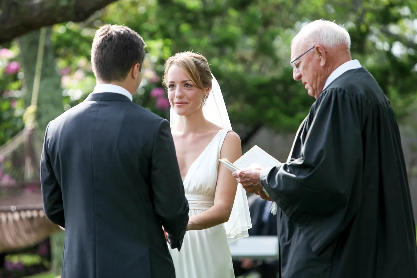 The officiant in a black robe reads vows as the bride looks up at the groom