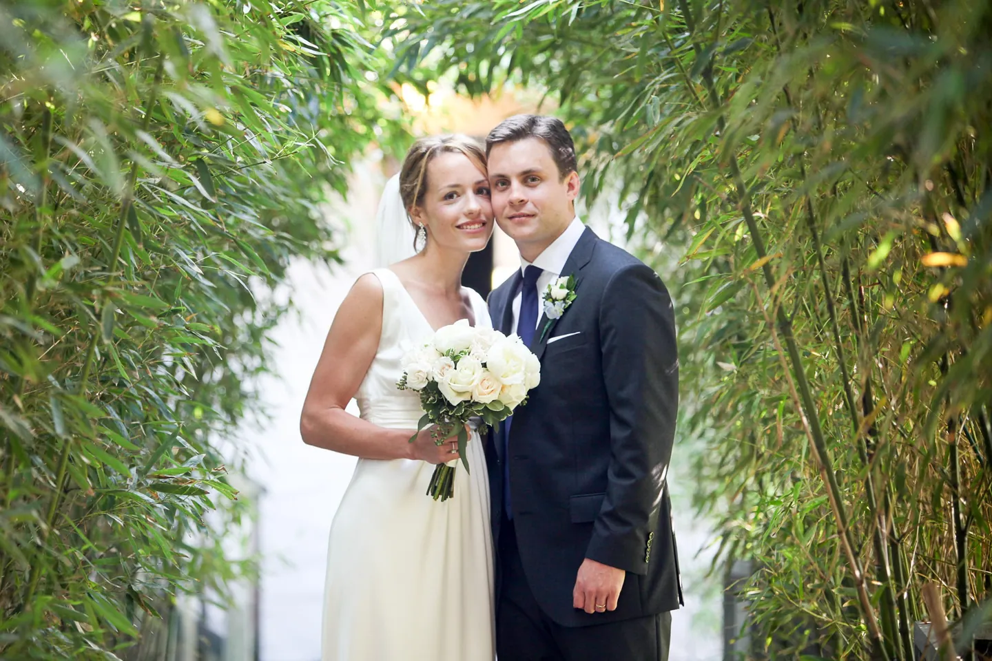 Kyla and Caley stand close in the bamboo corridor, her white bouquet between them