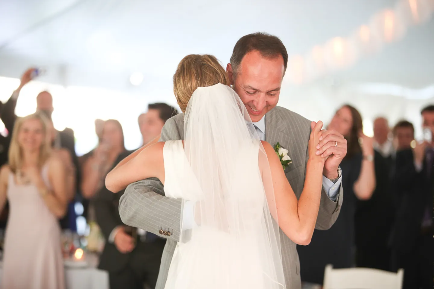 The bride dances with an older man in grey as guests clap under the tent