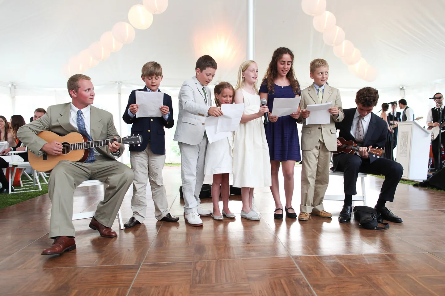 Children in blazers and dresses sing from lyric sheets as 2 men play guitar and ukulele