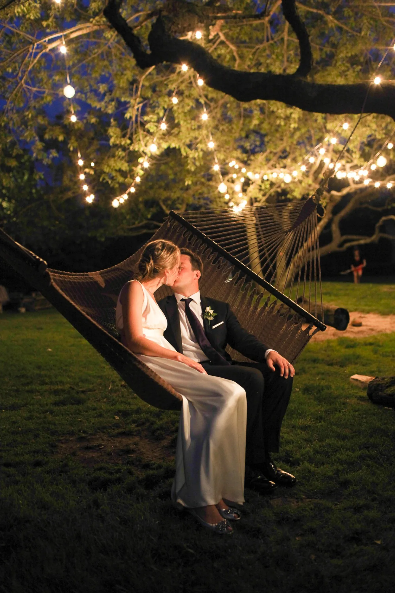 Kyla and Caley kiss in a rope hammock beneath a tree strung with warm lights