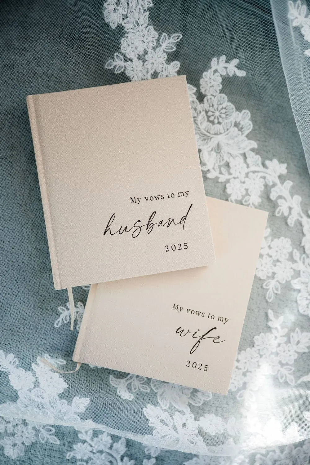 Two cream vow books, one for a husband and one for a wife, resting on lace