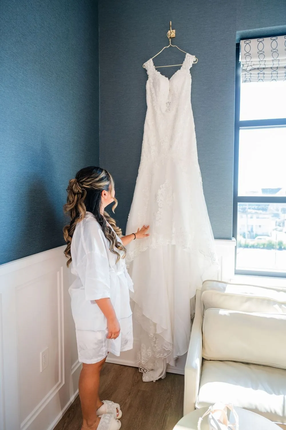 Wedding gown hangs in a bright corner by the window as the bride looks up at it