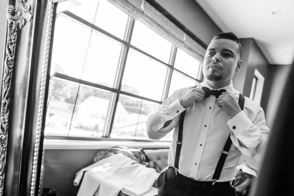 Black and white frame of the groom tying his bow tie by a tall window