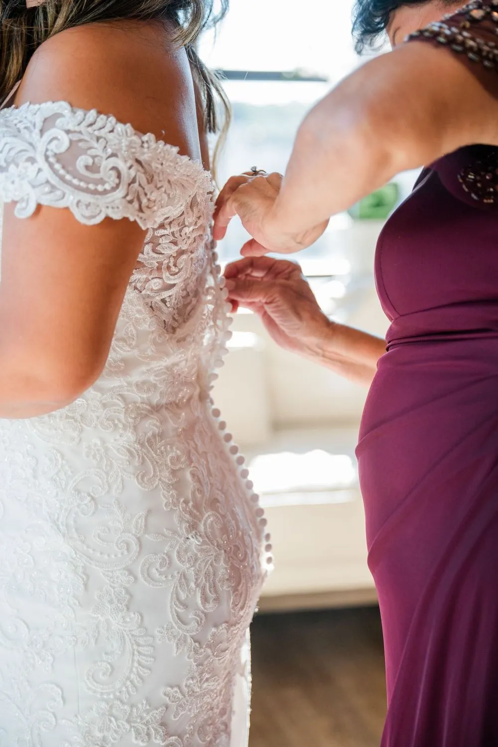 A woman in burgundy fastens the lace buttons down the back of the bride's gown