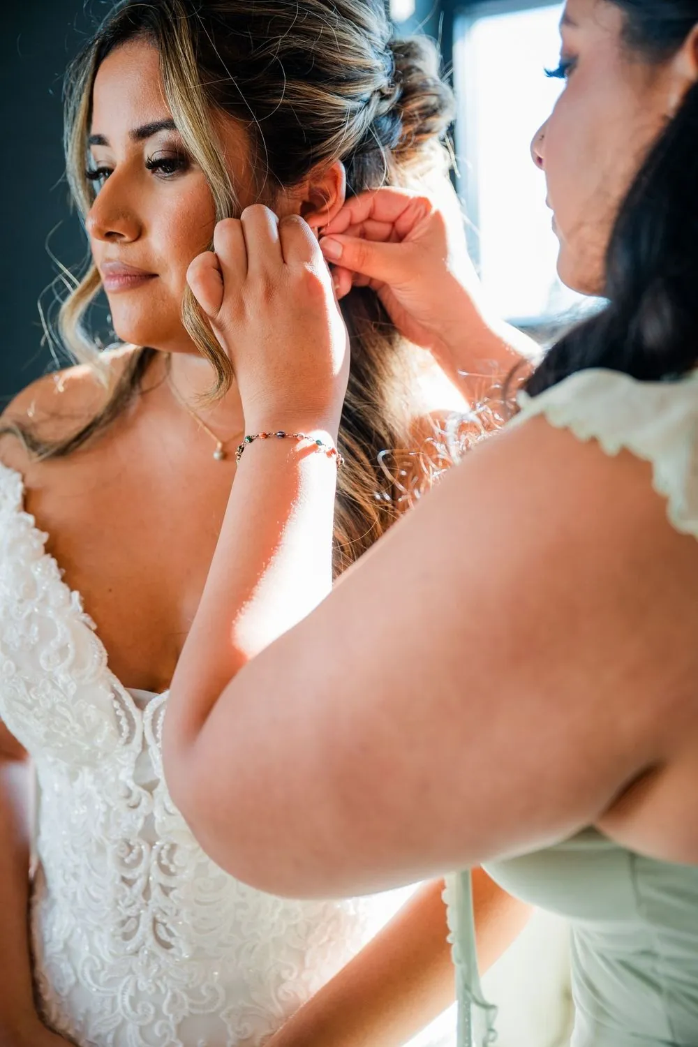 Bride puts in her earrings as a bridesmaid helps adjust her hair by the window