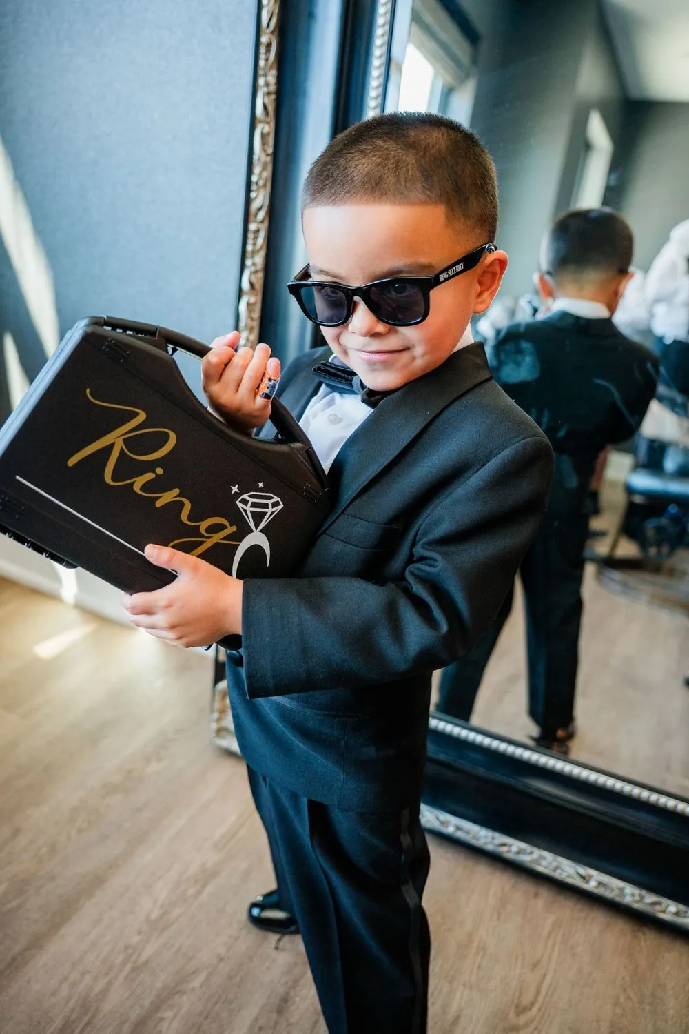 Ring bearer in sunglasses and a tux holds a black ring box sign