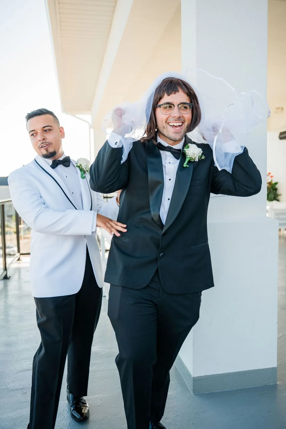 Groom in a white dinner jacket laughs as the best man lifts the veil off his wig