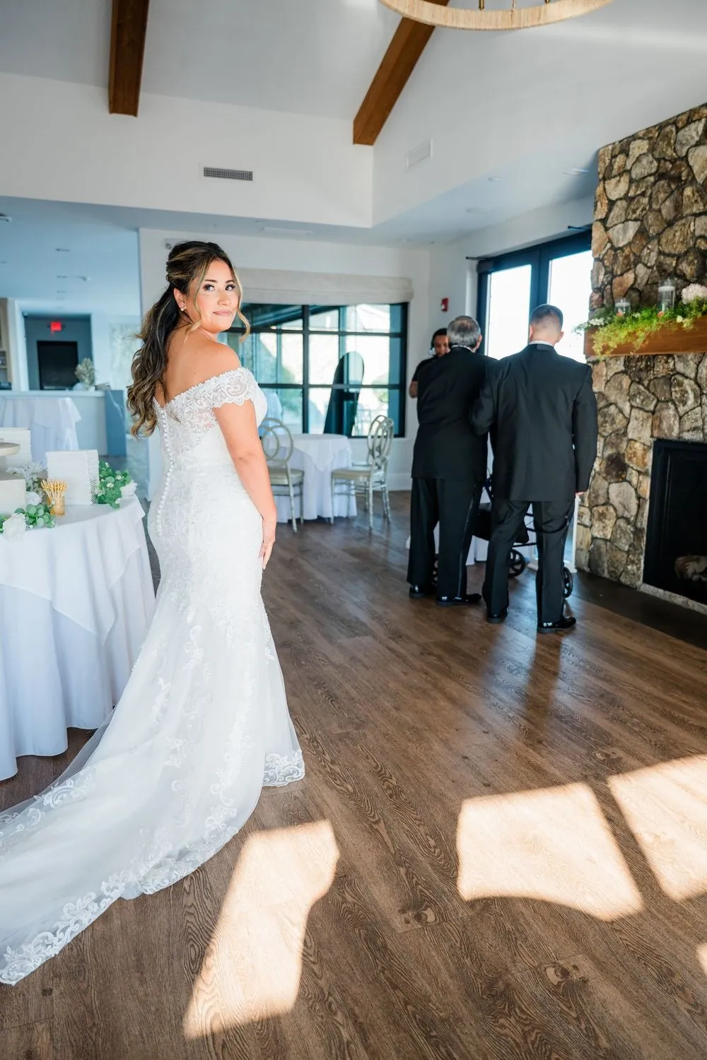 Bride in her gown walks toward two men waiting indoors by a stone fireplace