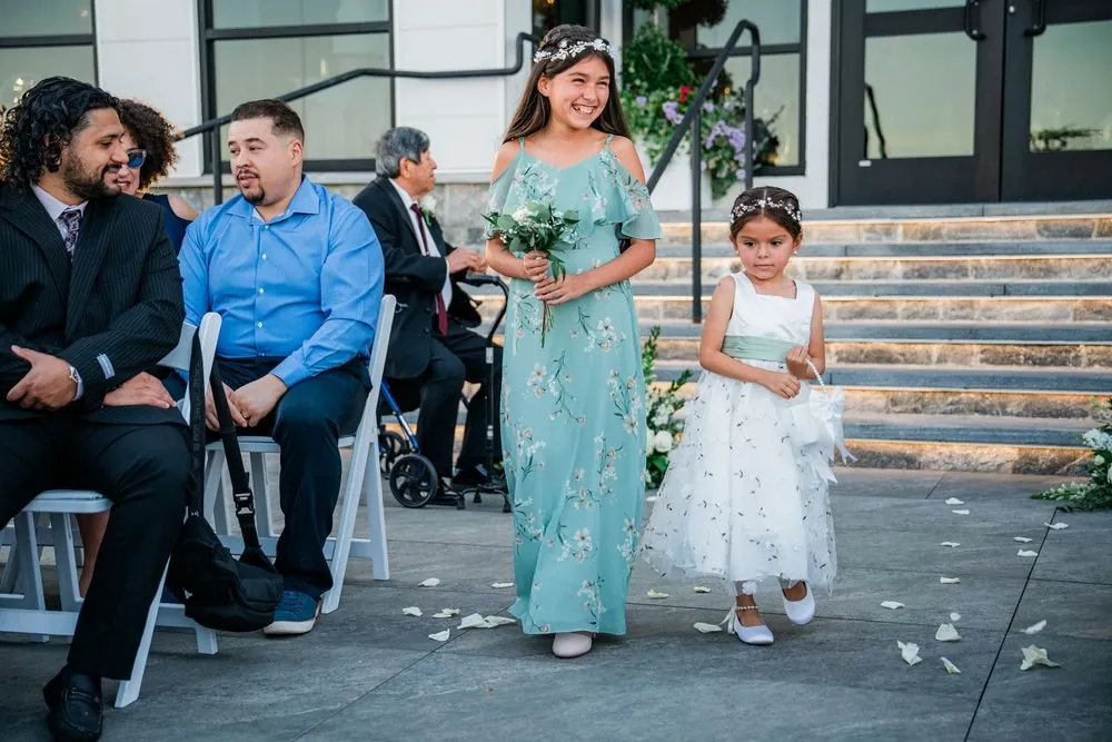Flower girl scatters petals down the terrace aisle beside a junior bridesmaid in sage