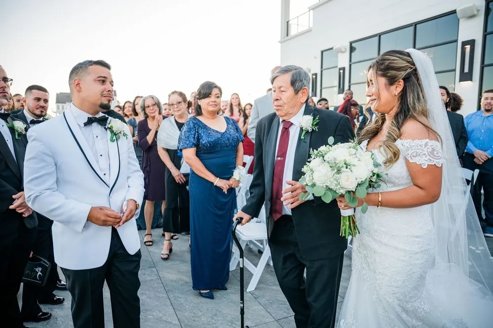Leslie's father walks her down the aisle as Miguel waits in a white dinner jacket