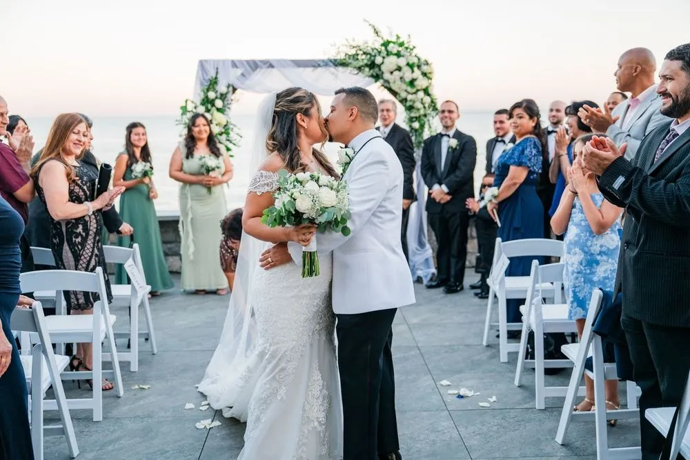 Leslie and Miguel kiss in the aisle as guests stand and applaud on the terrace