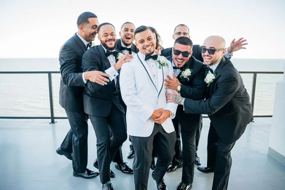 Groomsmen in black tuxedos crowd around the laughing groom on the balcony above the water