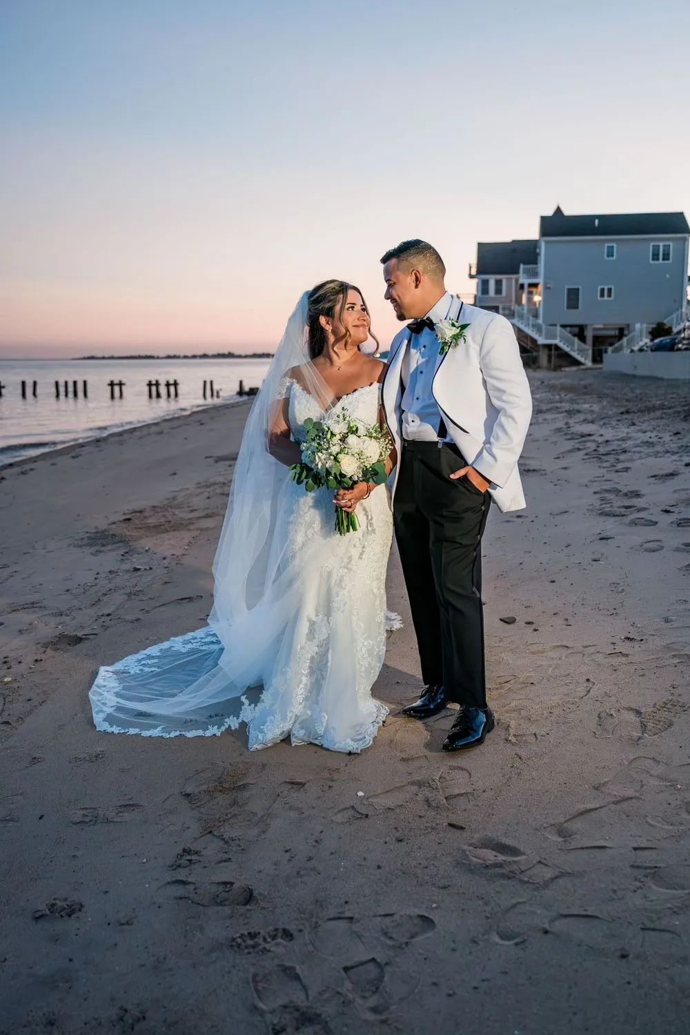 Leslie and Miguel stand close on the beach at dusk with houses on stilts behind them