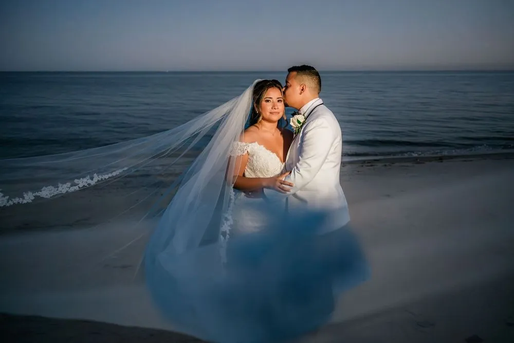 Miguel kisses Leslie's cheek on the beach at blue hour, her long veil drifting over the sand