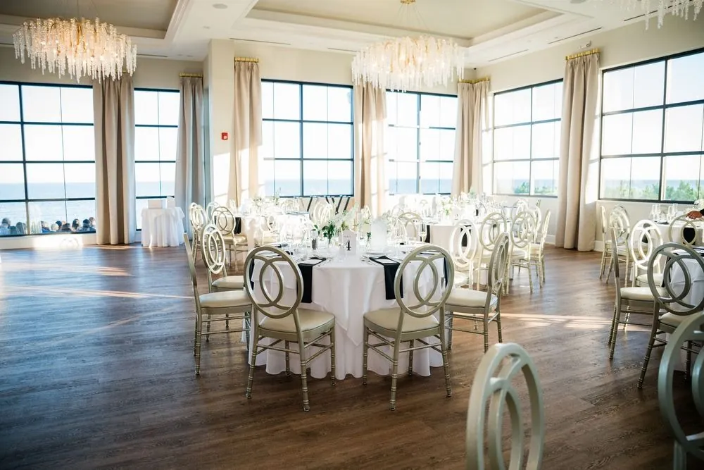 Reception room with white linens, gold chairs, and tall windows toward the beach