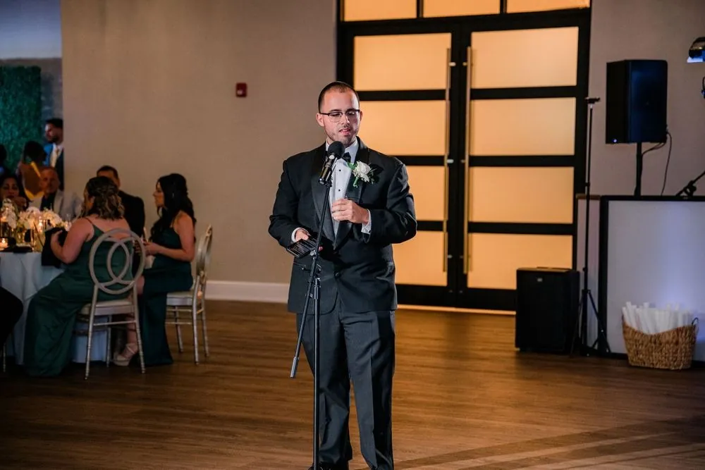 Guest in a black tuxedo gives a toast at a microphone stand in the dim reception room