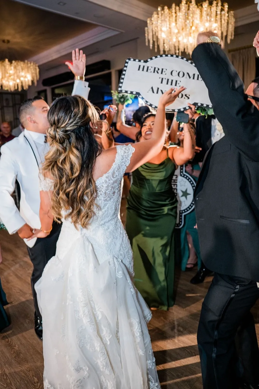 Bride dances into the room under chandeliers as guests wave a handmade sign