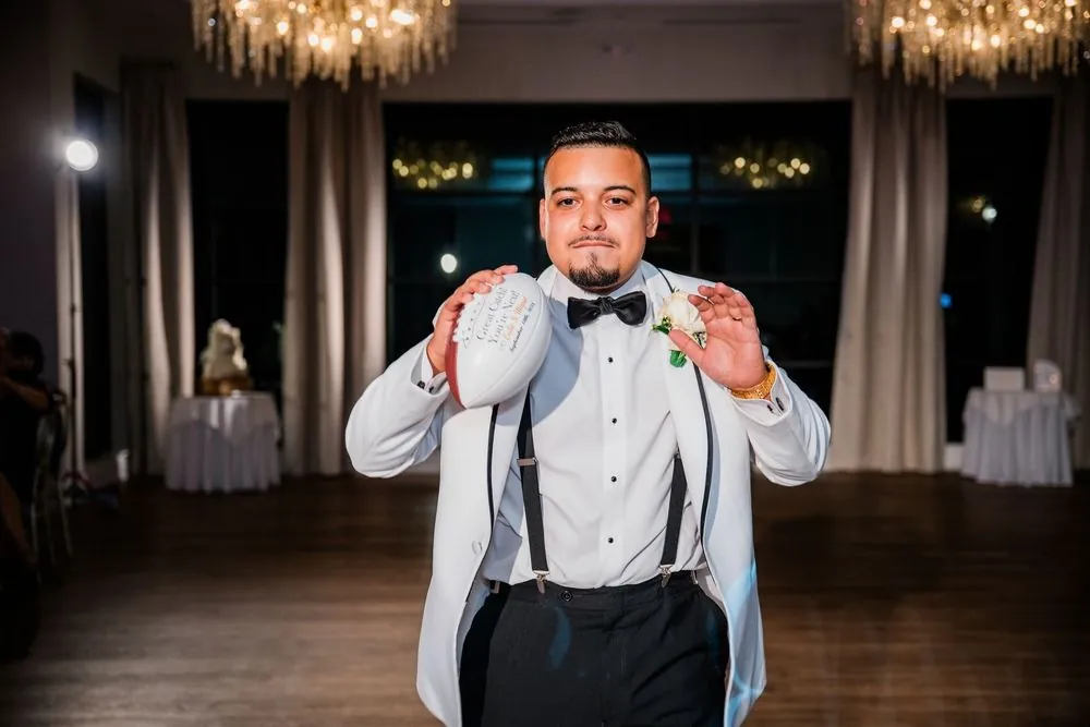 Groom holds up a white signed football on the dance floor under chandeliers
