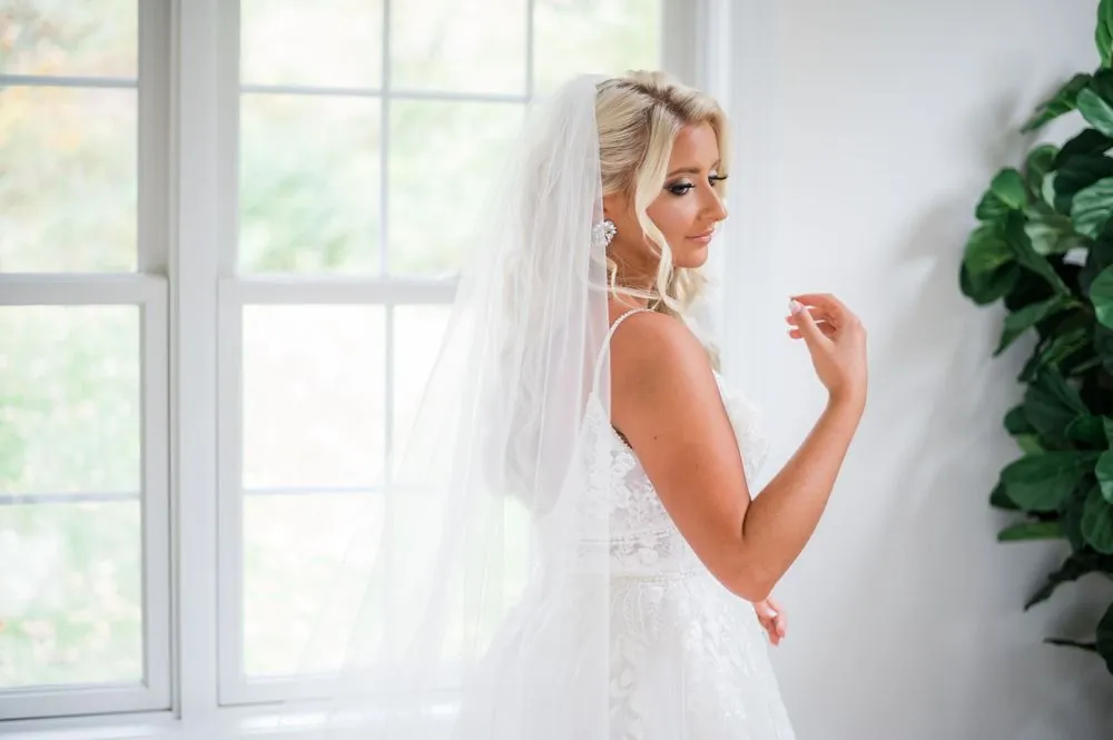 The bride adjusts her veil beside a tall bright window, lace straps catching the light