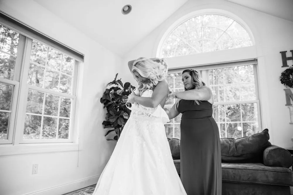 A bridesmaid laces up the back of the bride's gown under an arched window, in black and white