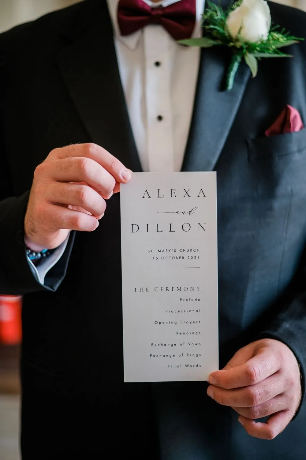 The groom holds a ceremony program printed with the couple's names against his tuxedo