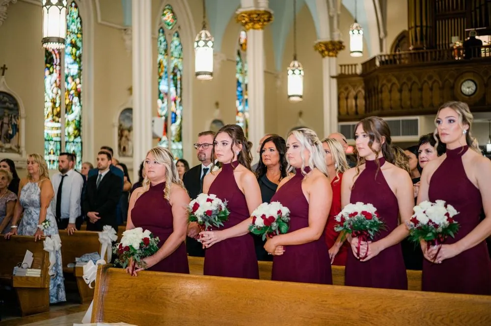 Bridesmaids in burgundy gowns watch the ceremony from the front pews below stained glass