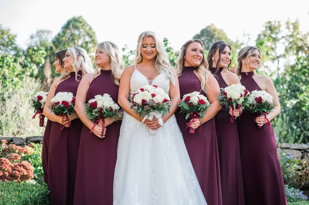 The bride smiles among bridesmaids in burgundy halter gowns holding red and white bouquets