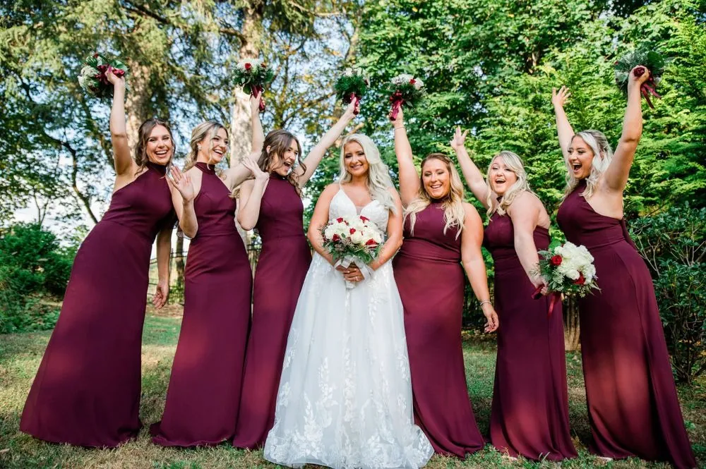 Bridesmaids in burgundy throw their arms up around the bride on a green lawn
