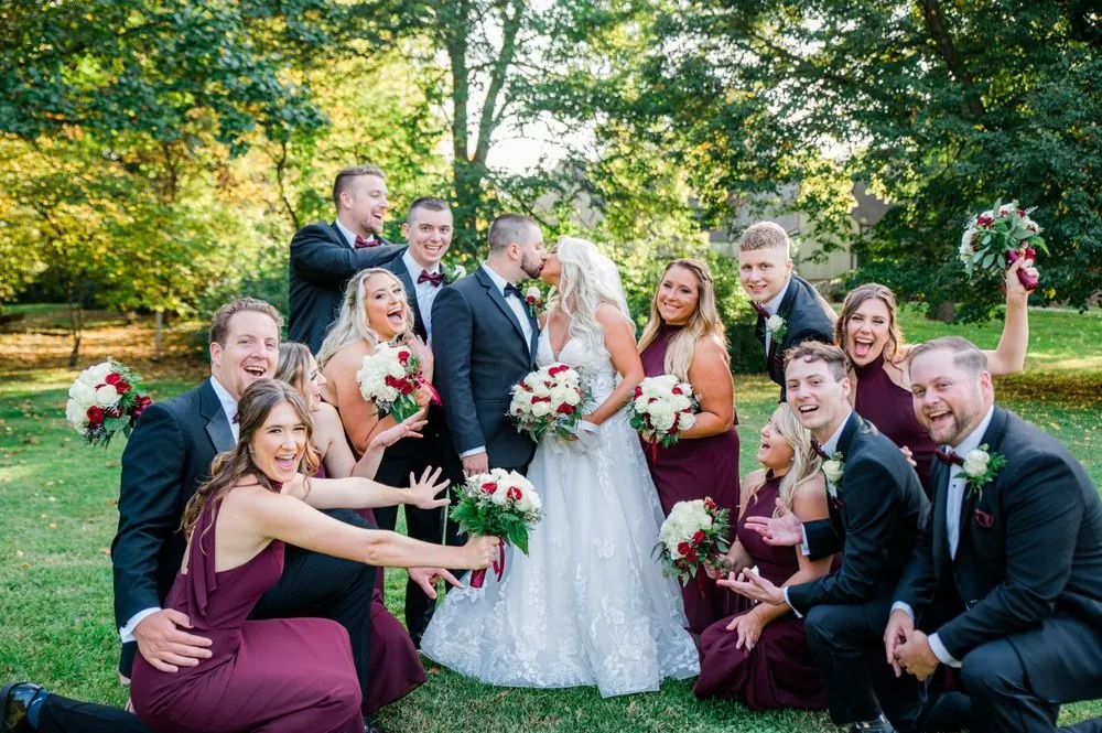 The full wedding party cheers and laughs on the lawn beneath tall trees
