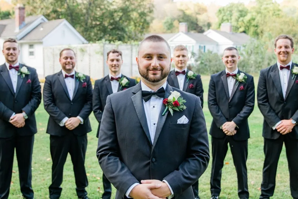 The groom stands in front of his groomsmen in black tuxedos on a sunny lawn