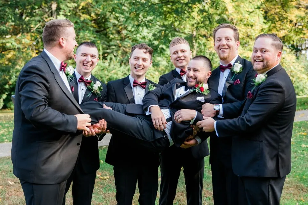 Groomsmen hoist the laughing groom sideways into the air on the grass