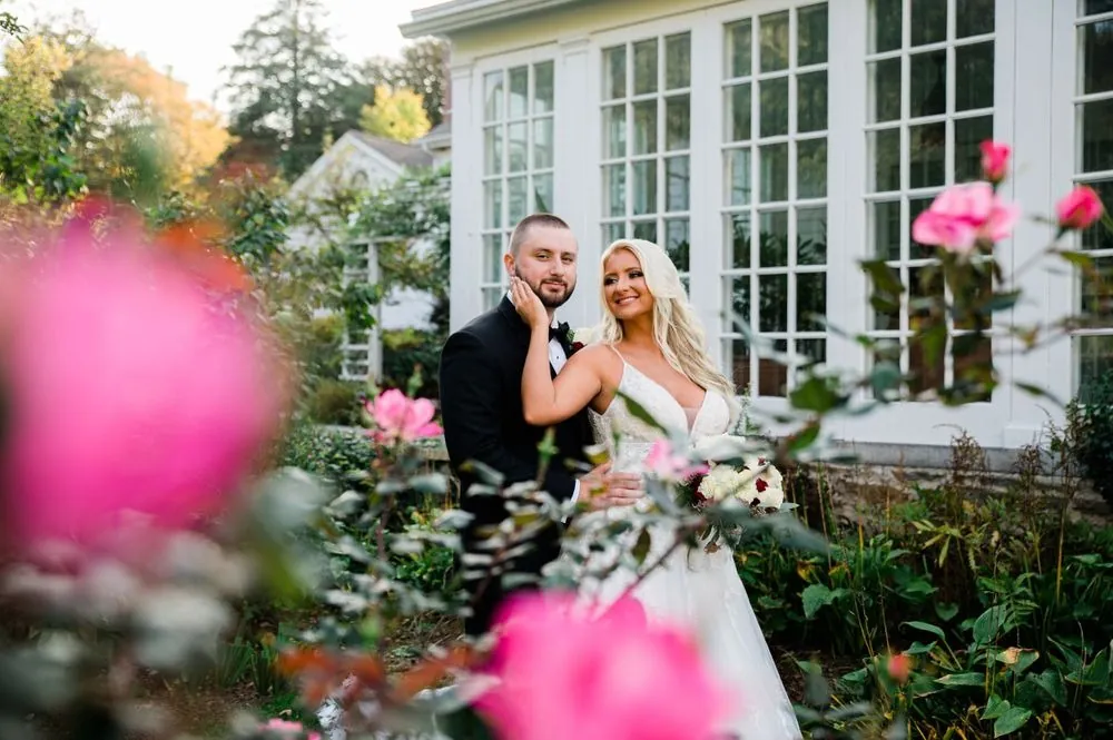 Lexy and Dillon smile beside tall windows, framed by blurred pink roses