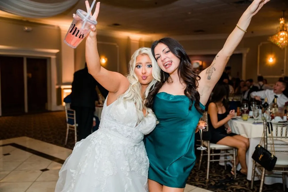 The bride and a friend in green throw their arms up at the reception