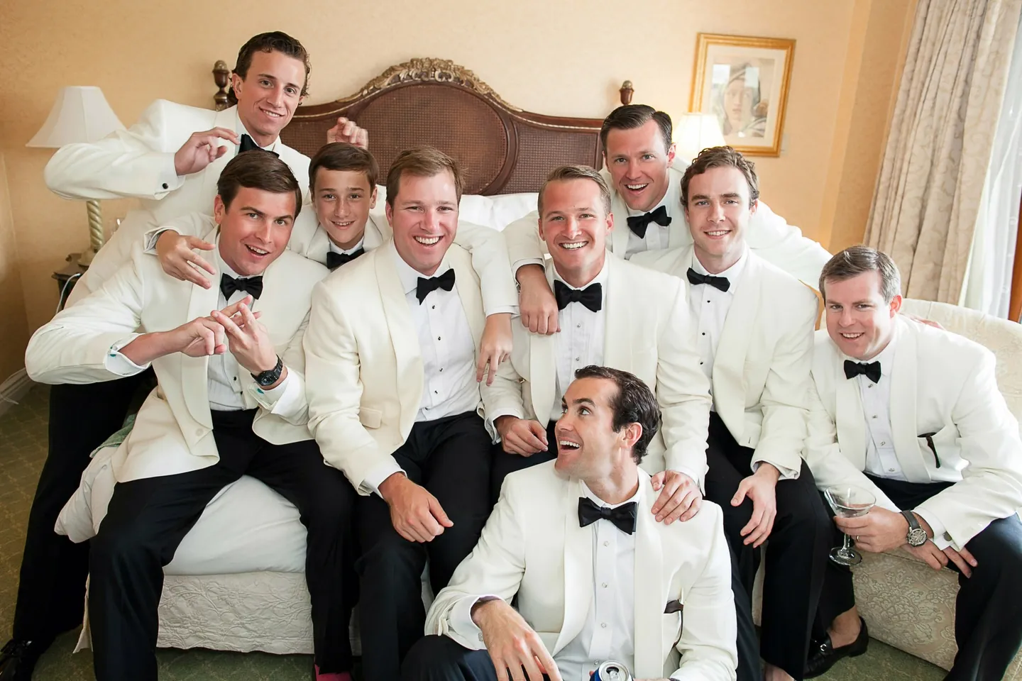 Groomsmen in white dinner jackets pile onto a sofa and laugh together