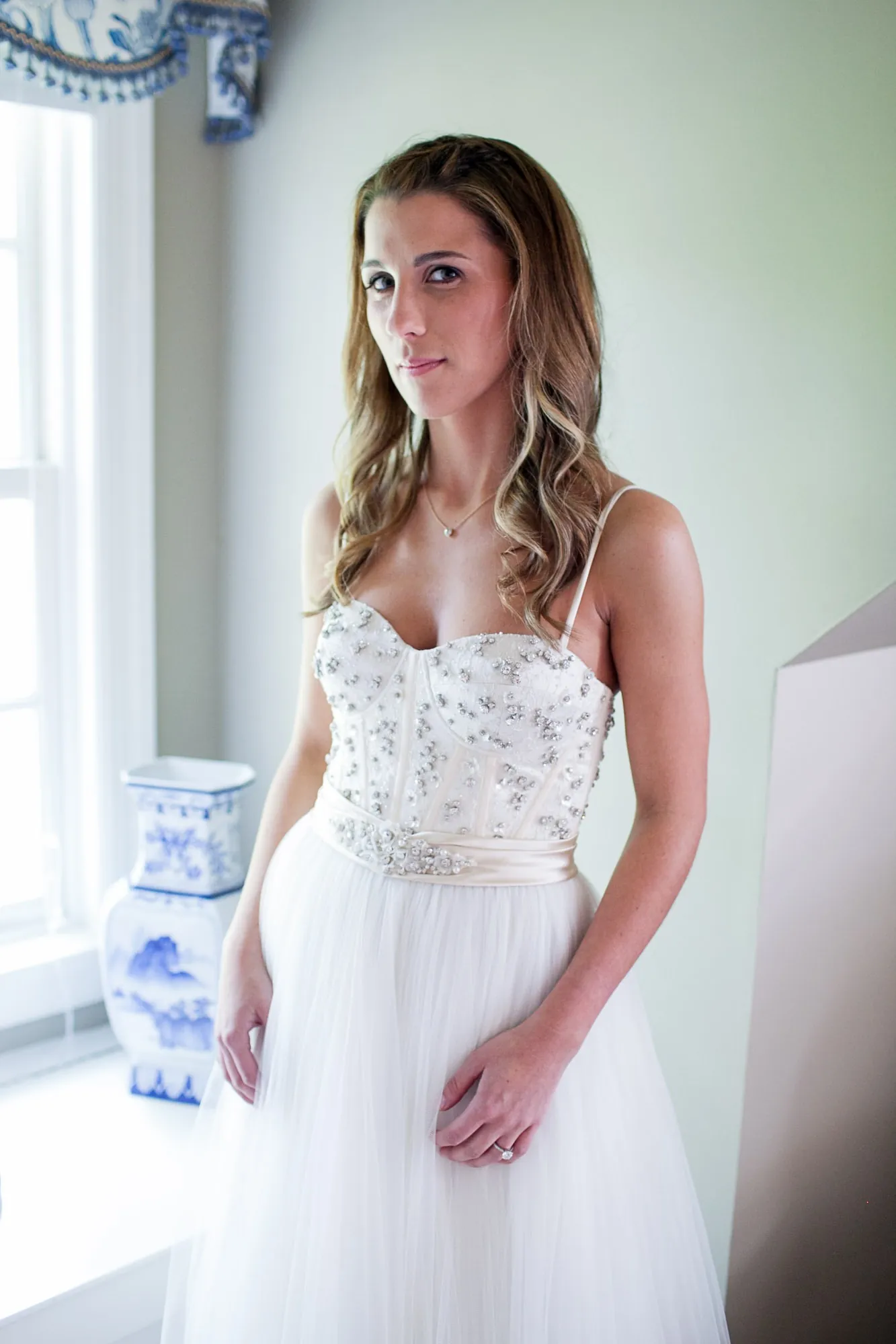The bride stands by a bright window in a beaded strapless gown, hair loose