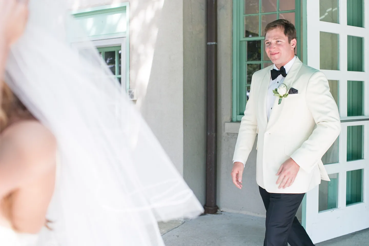 The groom in a white dinner jacket steps through a door as the bride's veil blows into the frame