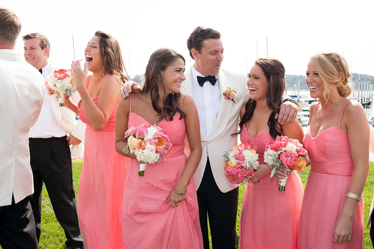 Bridesmaids in salmon gowns laugh with the groom near the marina