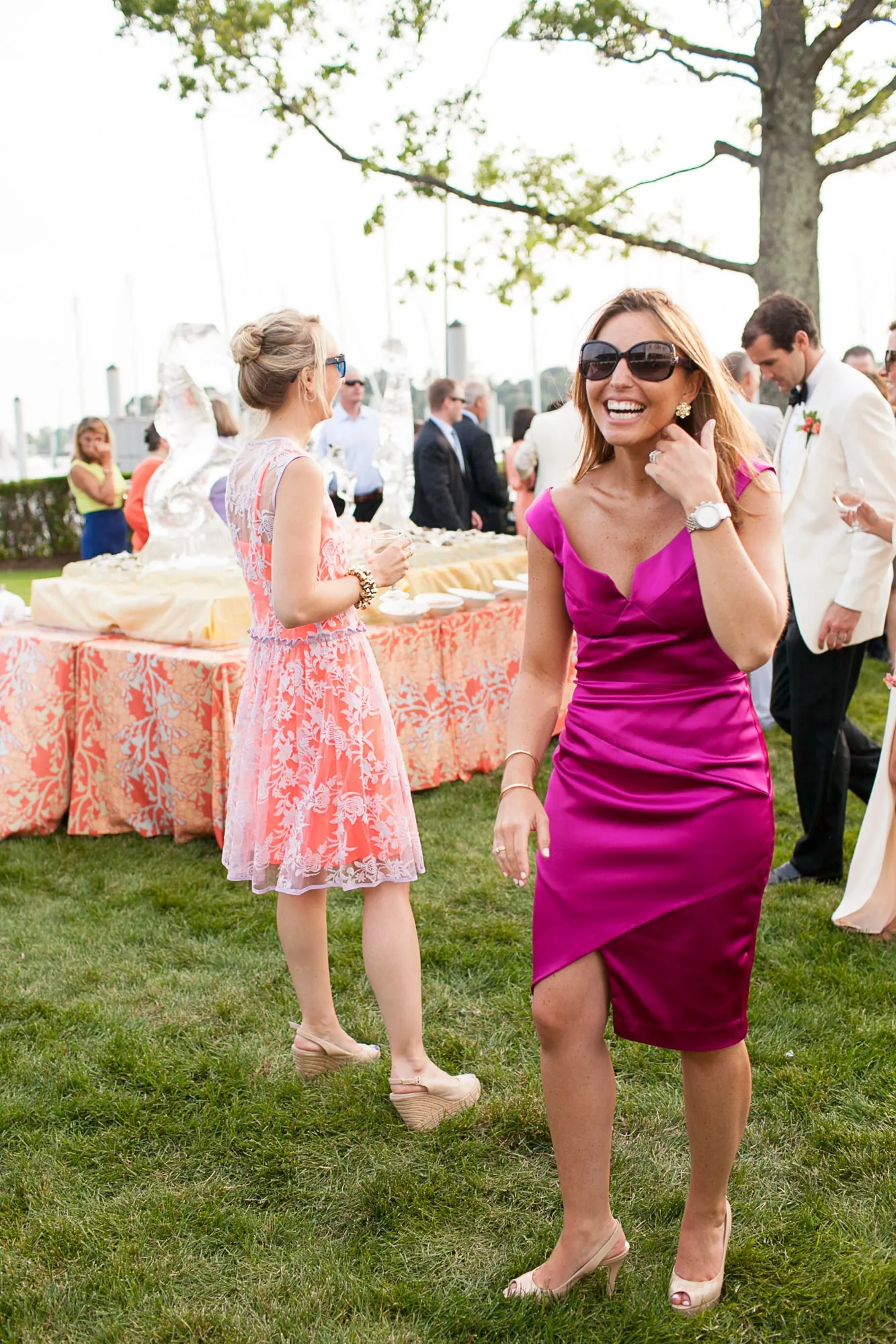 A guest in a magenta dress and sunglasses laughs on the lawn during cocktail hour
