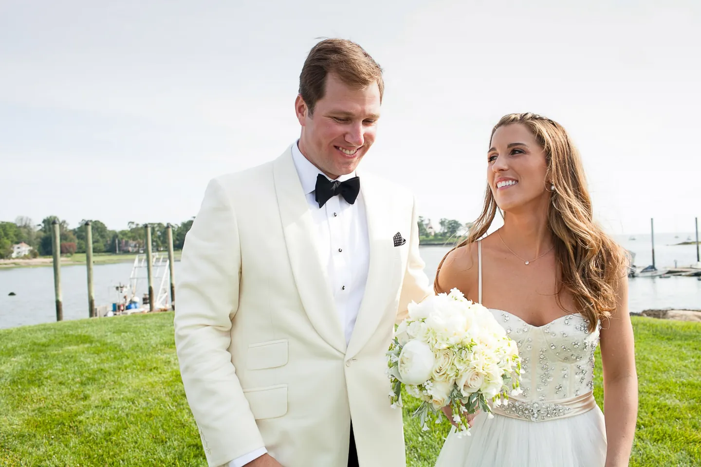 Maddie and Dave smile side by side on the lawn with boats on the water behind