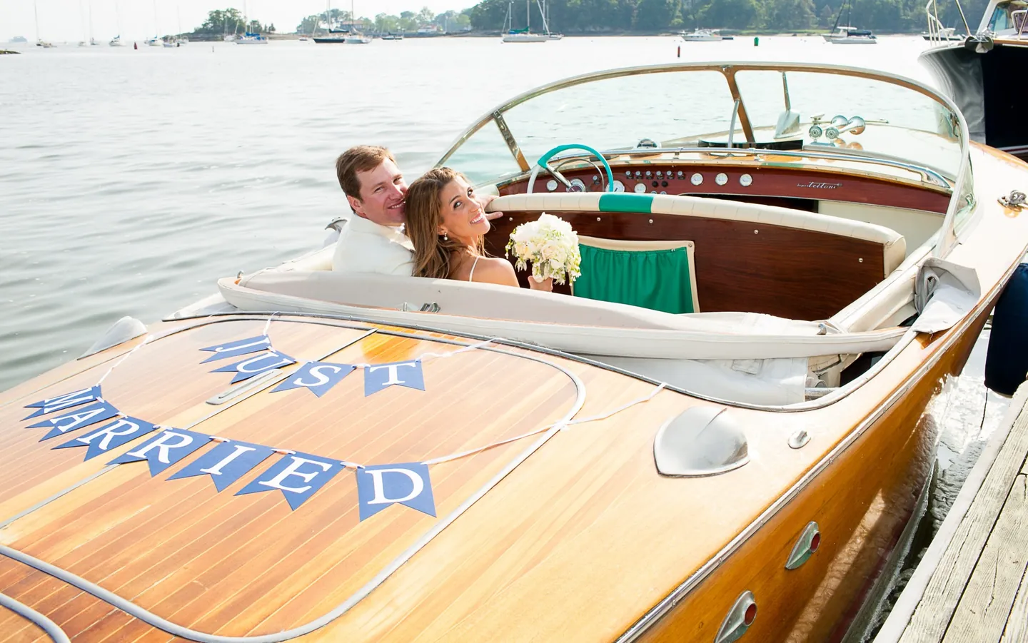 Maddie and Dave sit in a varnished wooden runabout with a Just Married banner on the deck