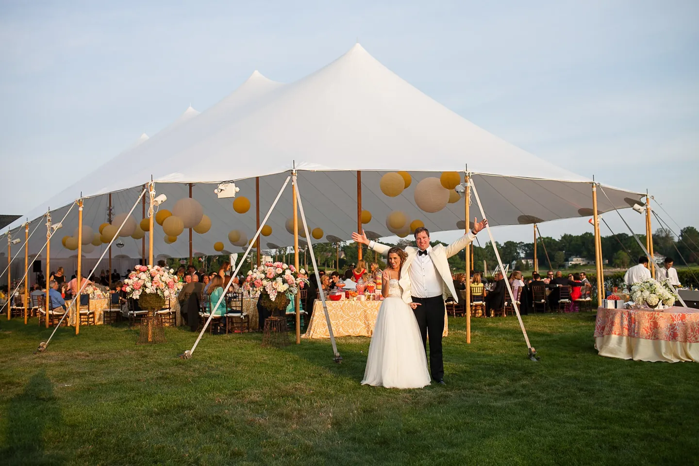 Maddie and Dave throw their arms up on the lawn in front of the lantern-filled sailcloth tent