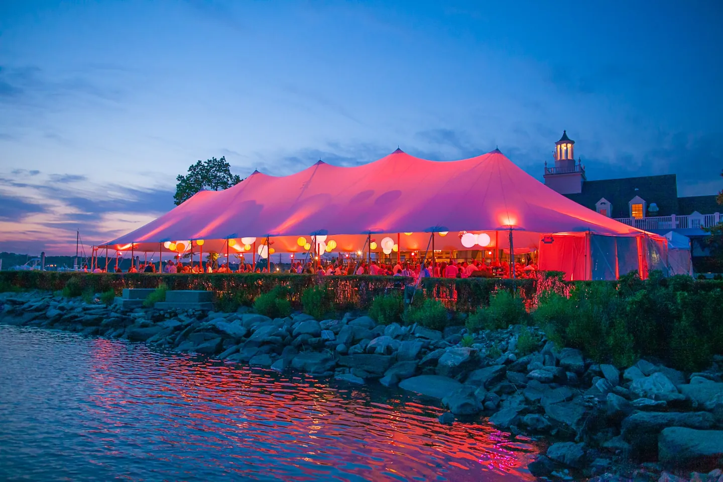 The pink-lit tent reflected across the harbor beyond a rocky seawall at dusk