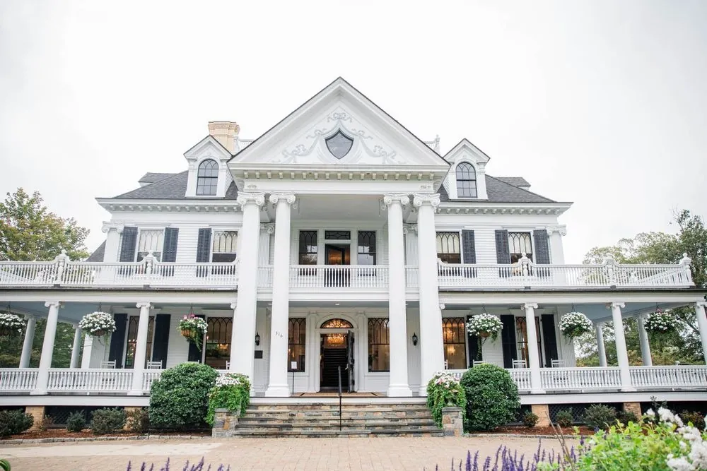 The white Lounsbury House with its wraparound porch and columns under an overcast sky