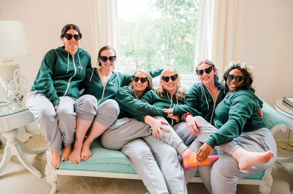 Bridesmaids in matching green hoodies and sunglasses pose together on a white sofa