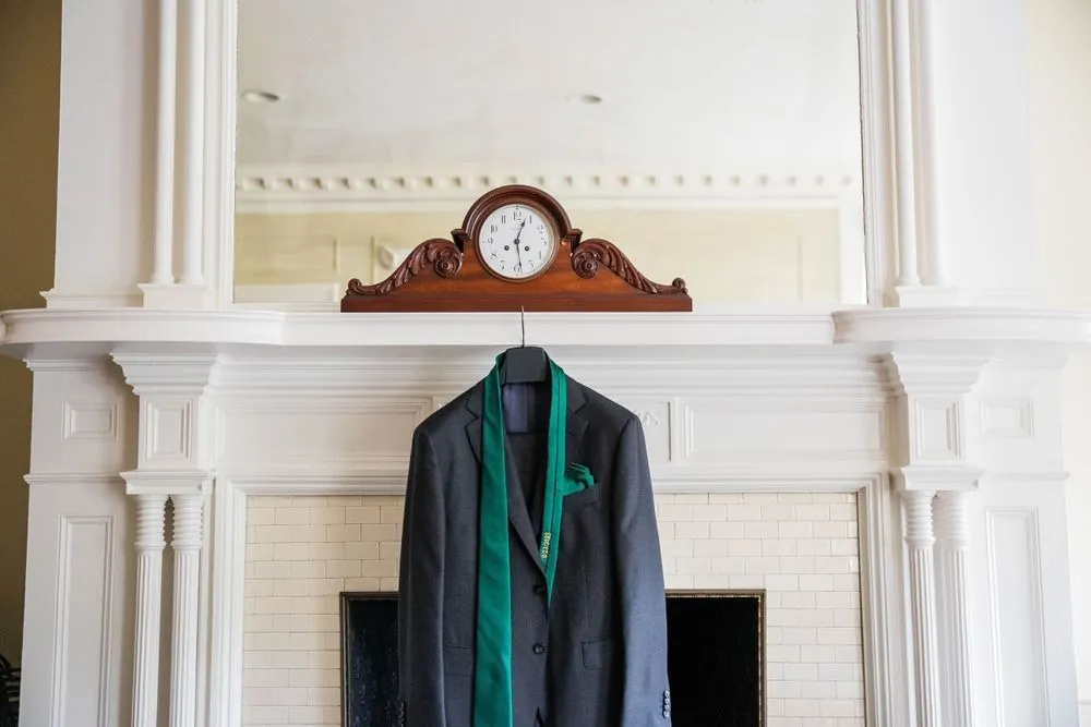The groom's gray suit and green tie hang from a white mantel beneath a carved clock