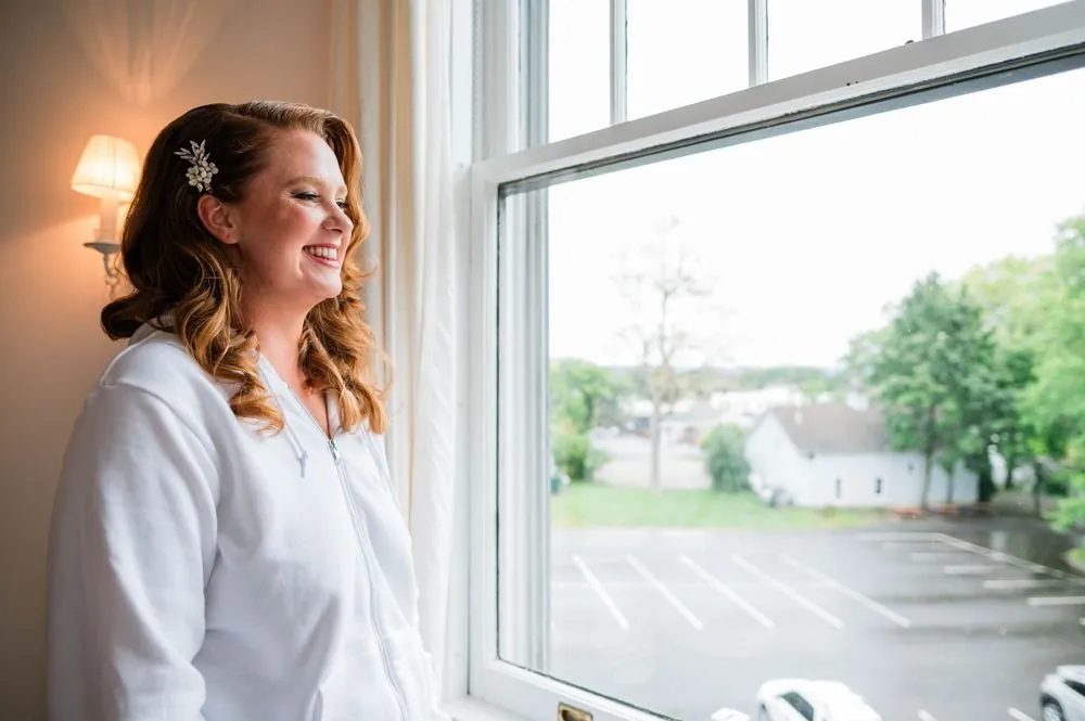 The bride smiles beside a rain-streaked window with a pearl comb in her red curls
