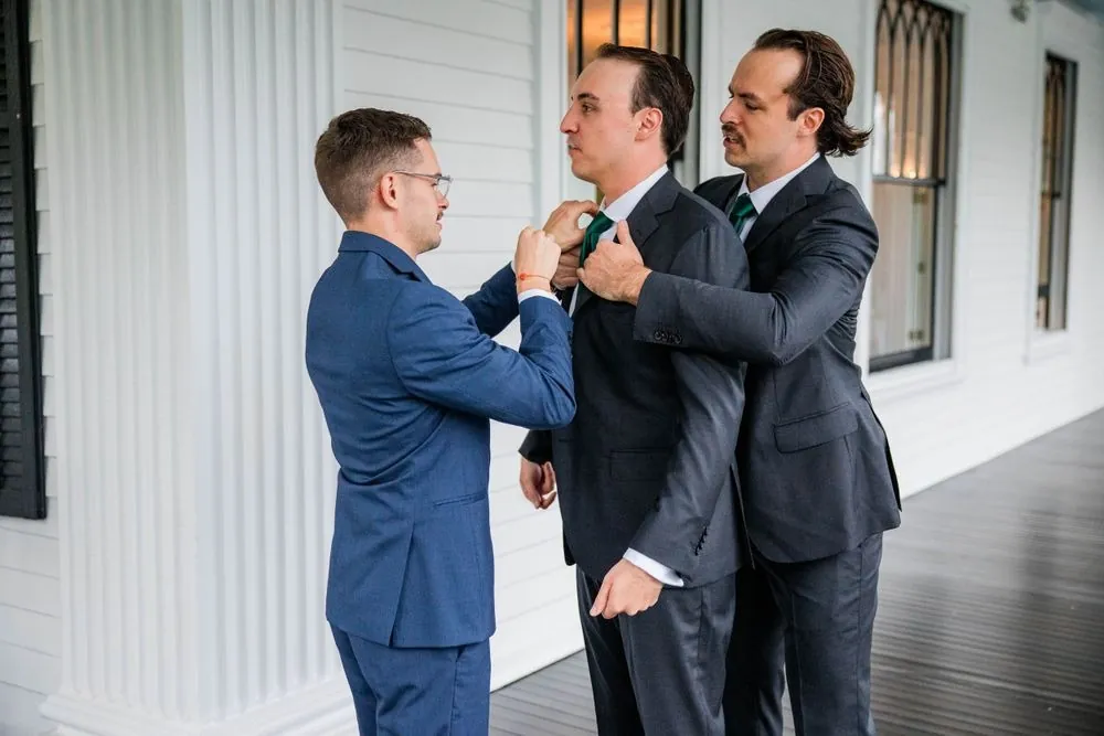 Groomsmen help pin a boutonniere on the groom in a white-paneled room